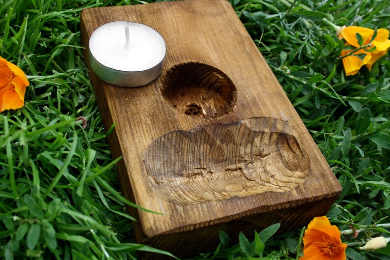 May include: A rustic, wooden candle holder with a natural finish. It features a circular indentation for a tea light candle and a carved, organic shape. The candle holder is set on green grass with orange flowers.