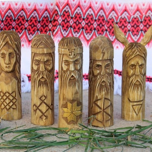 May include: Five wooden figurines carved with faces and symbols. The figurines are arranged in a row on a brown surface with green grass and pine cones.