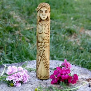 May include: A wooden carving of a female figure with intricate details and a carved inscription at the bottom that reads "Синяя". The carving is standing on a tree stump with pink flowers.