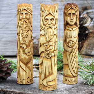 May include: Three wooden carvings of deities, each with a bird in their hands. The carvings are detailed and feature the names "Dian Ces", "Cernunnos", and "Anghb", carved at the bottom.