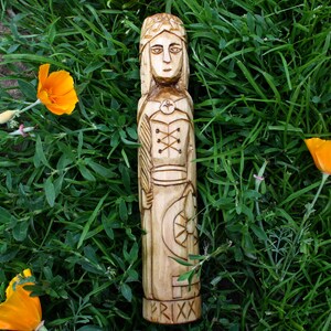 May include: A carved wooden figure of a woman with a crown, standing on a bed of green grass with orange flowers. The figure has detailed carvings of clothing and a shield. The base of the figure has the word "FRIXX" carved into it.