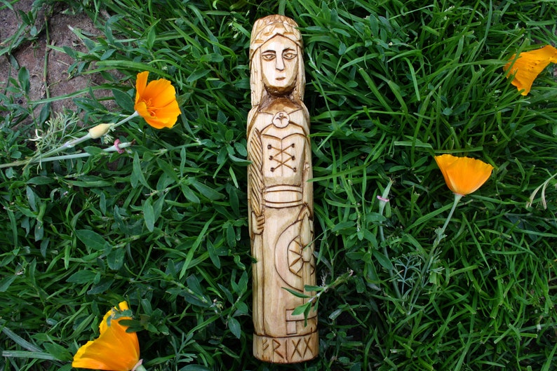 May include: A carved wooden figure of a woman with a crown, standing on a bed of green grass with orange flowers. The figure has detailed carvings of clothing and a shield. The base of the figure has the word "FRIXX" carved into it.