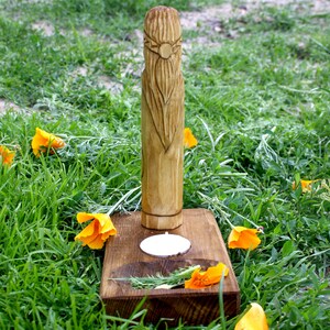 May include: A carved wooden figure with a long beard and a sun-like design on its head, standing on a wooden base with a small candle holder. The base is surrounded by orange flowers and green grass.