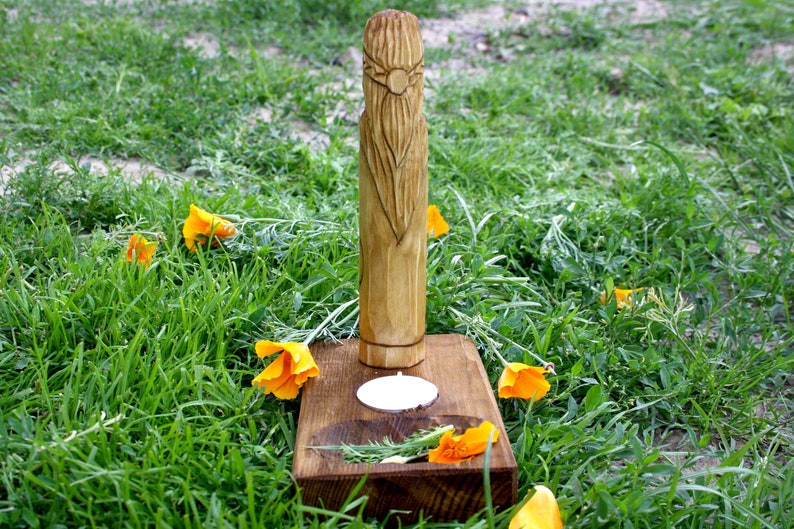 May include: A carved wooden figure with a long beard and a sun-like design on its head, standing on a wooden base with a small candle holder. The base is surrounded by orange flowers and green grass.