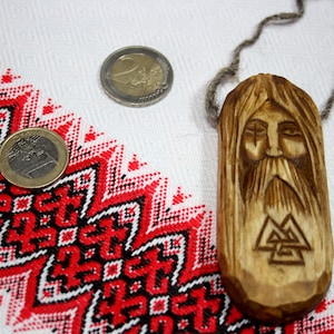 May include: A wooden pendant featuring a carved face and a triple triangle symbol. The pendant is displayed against a white background with a red and black pattern.