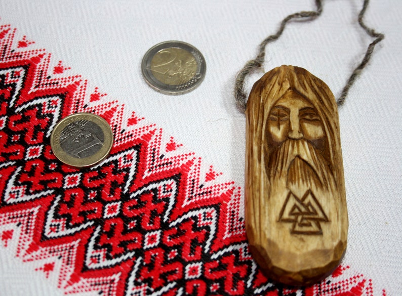 May include: A wooden pendant featuring a carved face and a triple triangle symbol. The pendant is displayed against a white background with a red and black pattern.