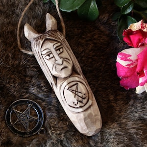 Puede incluir: Un colgante de madera tallada con una cara y cuernos, con un símbolo. También hay un amuleto de pentagrama de metal. Los artículos descansan sobre una superficie similar a la piel, con una rosa rosa y blanca en el fondo.