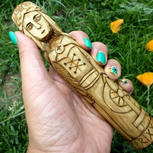 May include: A hand-carved wooden figurine of a woman with intricate details. The figure is light brown with carved facial features, clothing, and decorative patterns. The figurine is held against a backdrop of green grass and yellow flowers.