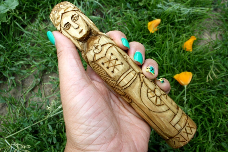 May include: A hand-carved wooden figurine of a woman with intricate details. The figure is light brown with carved facial features, clothing, and decorative patterns. The figurine is held against a backdrop of green grass and yellow flowers.