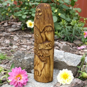 K&ouml;nnte beinhalten: Eine Holzschnitzerei einer b&auml;rtigen Figur, die einen Stab h&auml;lt und auf einem Bett aus braunen Holzsp&auml;nen und Steinen steht. Die Figur ist im rustikalen Stil geschnitzt mit Details von Kleidung und einem Bart.