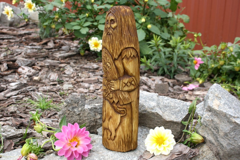 K&ouml;nnte beinhalten: Eine Holzschnitzerei einer b&auml;rtigen Figur, die einen Stab h&auml;lt und auf einem Bett aus braunen Holzsp&auml;nen und Steinen steht. Die Figur ist im rustikalen Stil geschnitzt mit Details von Kleidung und einem Bart.