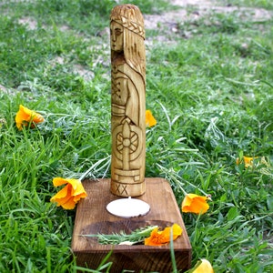 May include: A carved wooden statue of a woman with a crown, standing on a wooden base with a candle. The statue is surrounded by green grass and orange flowers. The statue is approximately 12 inches tall.