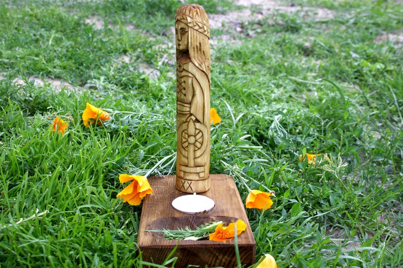 May include: A carved wooden statue of a woman with a crown, standing on a wooden base with a candle. The statue is surrounded by green grass and orange flowers. The statue is approximately 12 inches tall.