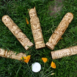 May include: Five intricately carved wooden figures, each depicting a different Viking-era character, are arranged on a bed of green grass. The figures are light brown with detailed carvings, and some have helmets or other adornments. A small candle and orange flowers are also present.