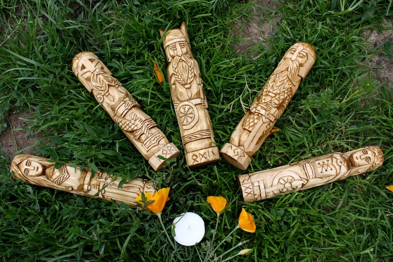 May include: Five intricately carved wooden figures, each depicting a different Viking-era character, are arranged on a bed of green grass. The figures are light brown with detailed carvings, and some have helmets or other adornments. A small candle and orange flowers are also present.