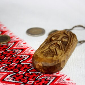 May include: A wooden pendant with a carved face and a braided cord necklace. The pendant is brown and has a detailed carving of a face with a beard. The necklace is made of a brown cord and is tied in a knot at the top.