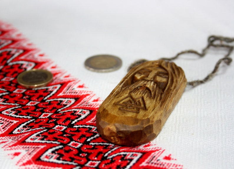 May include: A wooden pendant with a carved face and a braided cord necklace. The pendant is brown and has a detailed carving of a face with a beard. The necklace is made of a brown cord and is tied in a knot at the top.