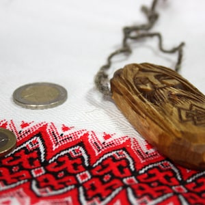 May include: A wooden pendant with a carved face and a braided cord. The pendant is lying on a white fabric with a red and black geometric pattern.