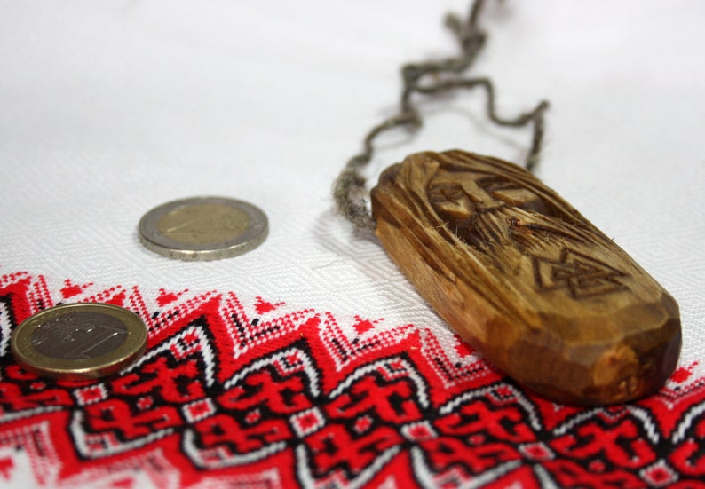 May include: A wooden pendant with a carved face and a braided cord. The pendant is lying on a white fabric with a red and black geometric pattern.