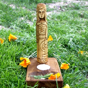 May include: A carved wooden statue of a figure with intricate details, standing on a wooden base with a candle. The statue is surrounded by green grass and bright orange flowers. The word "FRIXX" is carved on the base.