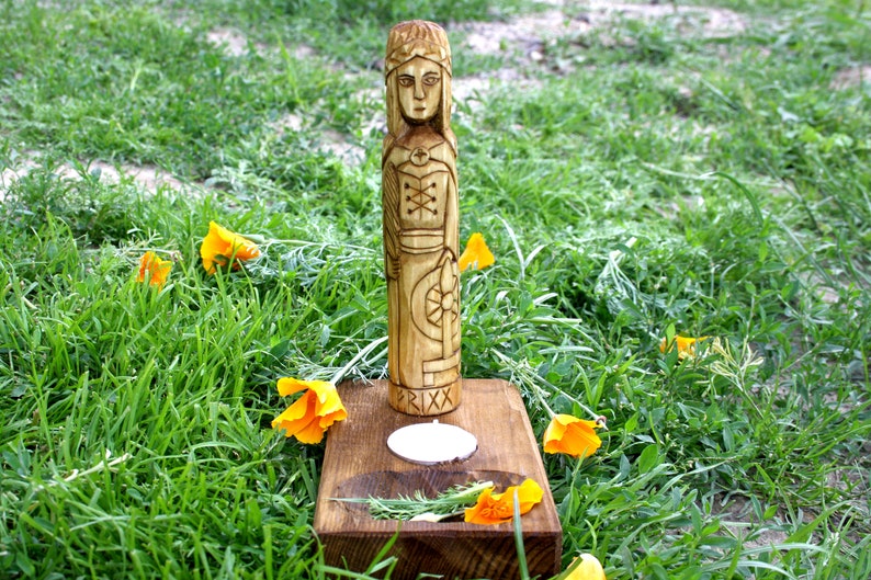 May include: A carved wooden statue of a figure with intricate details, standing on a wooden base with a candle. The statue is surrounded by green grass and bright orange flowers. The word "FRIXX" is carved on the base.