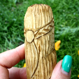 May include: A hand-carved wooden object with a detailed design. The object is cylindrical, with a textured surface and a carved band featuring a circular element and leaf-like details. The background is blurred green grass.
