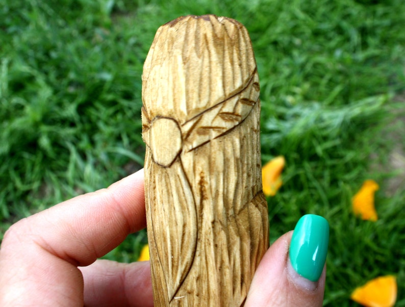 May include: A hand-carved wooden object with a detailed design. The object is cylindrical, with a textured surface and a carved band featuring a circular element and leaf-like details. The background is blurred green grass.