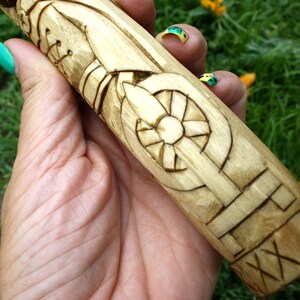 May include: A hand-carved wooden object with intricate geometric and stylized designs. The light-colored wood features detailed carvings of a wheel-like shape and other patterns. The object is held against a backdrop of green grass and yellow flower petals.