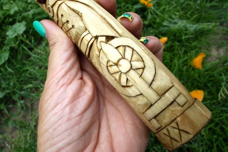 May include: A hand-carved wooden object with intricate geometric and stylized designs. The light-colored wood features detailed carvings of a wheel-like shape and other patterns. The object is held against a backdrop of green grass and yellow flower petals.