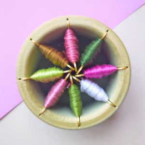 Puede incluir: Un soporte de madera para carretes con ocho carretes de hilo de bordar en varios colores, incluyendo rosa, verde, blanco y marrón.