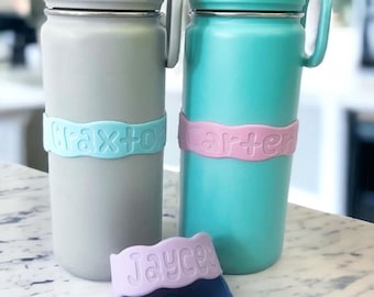May include: Two stainless steel water bottles with personalized labels. The bottle on the left has a light blue label with the name "Graxton" on it. The bottle on the right has a pink label with the name "Carter" on it. A purple label with the name "Jaycee" is in the foreground.