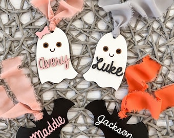 May include: Two white ghost ornaments with brown eyes and smiles, one featuring a pink bow and the name Avery, the other with the name Luke. Two black bat ornaments featuring the names Maddie in pink and Jackson in white, all with frayed ribbons.
