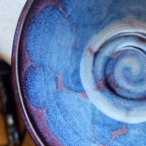 May include: A handmade ceramic bowl with a deep blue interior and a dark purple rim. The bowl's interior features a textured, cloud-like pattern with lighter blue and white swirls in the centre. The rim is a darker shade of purple.