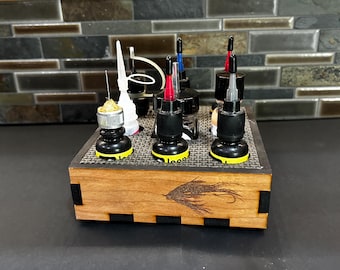 Pode incluir: Um organizador de madeira com várias garrafas e aplicadores, provavelmente para artesanato ou hobbies. O organizador tem um design de pesca com mosca gravado a laser na frente. As garrafas têm tampas pretas com pontas vermelhas, azuis e pretas.