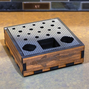 May include: A wooden box with a black and grey patterned interior. The box has a small compartment in the centre and several round holes around the edges. The bottom of the box is engraved with a fish.