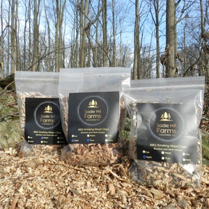 May include: Three clear plastic bags filled with wood chips for smoking meat. Each bag has a black label with the text "Sadie Hill Farms" and a list of wood types, including oak, apple, plum, cherry, and maple. The bags are sitting on a pile of wood chips.