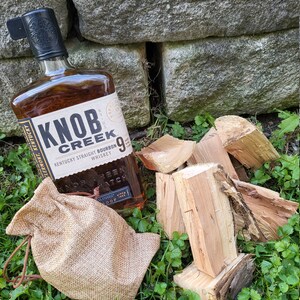 May include: A bottle of Knob Creek Kentucky Straight Bourbon Whiskey, with a brown burlap sack and pieces of firewood on a bed of green grass.