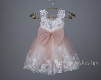 May include: A blush pink tulle dress with white lace detailing and a large blush pink bow at the back. The dress has a sleeveless design and a scalloped hem.