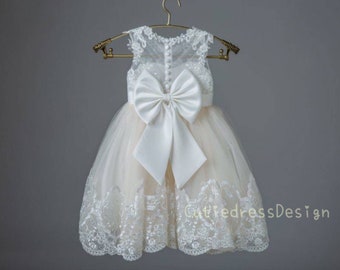May include: A white tulle dress with a lace overlay and a large white satin bow at the back. The dress has a sleeveless design and a round neckline.