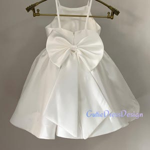 May include: A white satin dress with a large bow on the back, hanging on a gold-colored hanger. The dress has a halter neckline and a flared skirt. The text "CutieDressDesign" is visible on the lower right side of the dress.