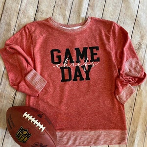May include: Red crew neck sweatshirt with the text "GAME DAY chiefs" in black and white. A brown football with white laces and the NFL logo is next to the sweatshirt. The sweatshirt has elbow patches and ribbed cuffs.
