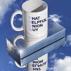 May include: White ceramic mug with a blue and black design that reads "That Helpful Union Guy". The mug is sitting on a silver surface with a reflection of the mug.