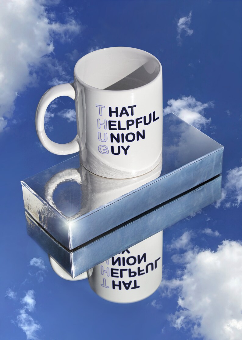 May include: White ceramic mug with a blue and black design that reads "That Helpful Union Guy". The mug is sitting on a silver surface with a reflection of the mug.