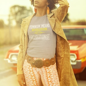 May include: A woman wearing a grey t-shirt with the text "Feminism Means Revolution and I am a Revolutionist" in yellow and white. She is wearing a brown suede jacket, a wide brown belt with a large metal buckle, and orange and white patterned pants. She is standing in front of a red car.