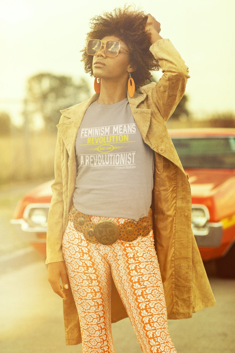 May include: A woman wearing a grey t-shirt with the text "Feminism Means Revolution and I am a Revolutionist" in yellow and white. She is wearing a brown suede jacket, a wide brown belt with a large metal buckle, and orange and white patterned pants. She is standing in front of a red car.