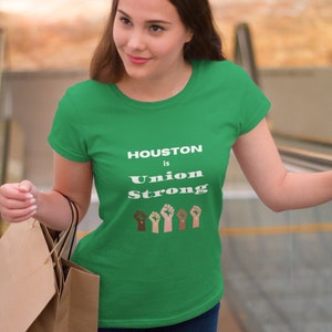 May include: A woman wearing a green t-shirt with the text "HOUSTON is Union Strong" and a row of raised fists in different skin tones. She is holding shopping bags.