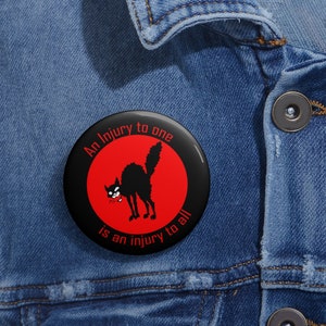 May include: A black and red button with the text "An injury to one is an injury to all" and a black cartoon cat with a red eye.