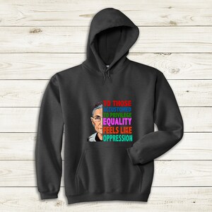 Op de afbeelding: Een donkergrijze hoodie met een kleurrijke afbeelding van een vrouwen gezicht en de tekst "Voor degenen die gewend zijn aan privileges, voelt gelijkheid als onderdrukking."