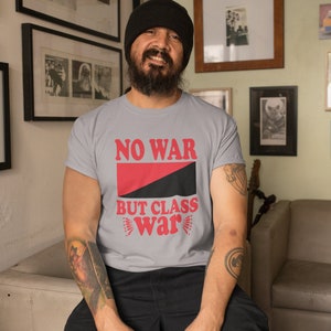 May include: A person wearing a grey t-shirt with the text "NO WAR BUT CLASS WAR" in red letters. The shirt features a black and red diagonal stripe. The person is wearing a black beanie and has a beard.