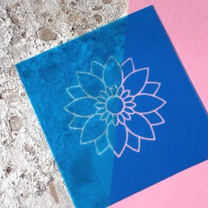 Könnte beinhalten: Eine quadratische, blaue Schablone mit einem weißen Blumendesign. Die Schablone befindet sich auf einer strukturierten, hellfarbigen Oberfläche und einer rosa Oberfläche. Das Blumendesign ist eine detaillierte Umrisszeichnung einer Blume.